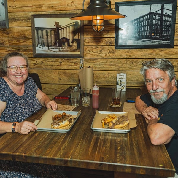 Two happy customers enjoying Inky's cheesesteaks and fries at a rustic wooden table.