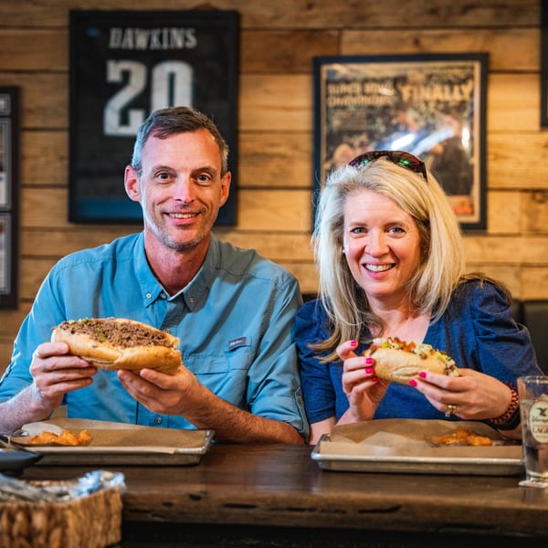 Smiling couple holding Inky's cheesesteaks, sitting at a wooden table with rustic decor.