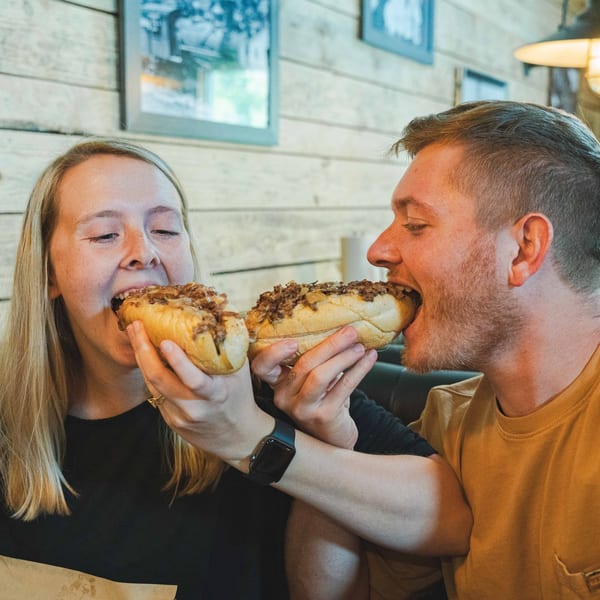 A couple enthusiastically eating Inky's cheesesteaks, with wooden walls and framed pictures in the background.