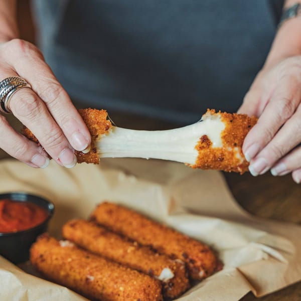 Hands pulling apart a mozzarella stick, revealing stretchy cheese, with more sticks and sauce below.