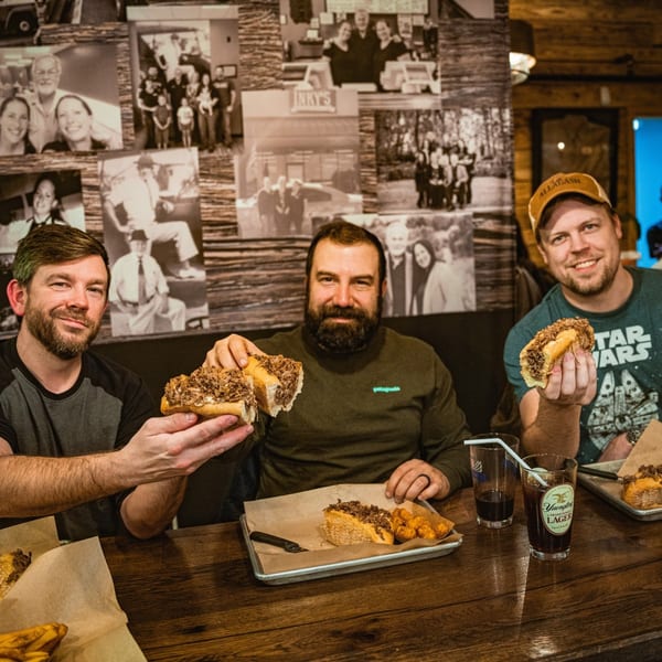 Three men smiling, holding up Inky's cheesesteaks, with family photos on the wall behind them.
