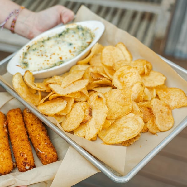 A tray of golden potato chips, a bowl of cheesy spinach dip, and mozzarella sticks.