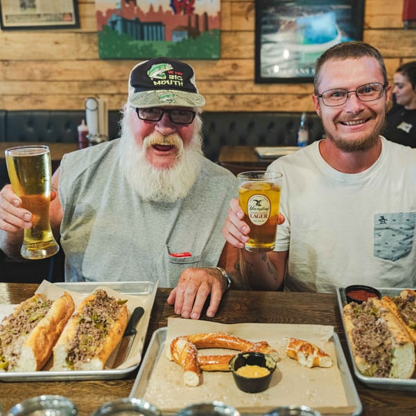 Two men smiling, holding beers, with Inky's cheesesteaks and pretzels on trays in front of them.
