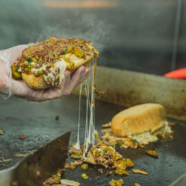 Gloved hands lift a steaming Inky's cheesesteak, cheese stretching, from a hot griddle.