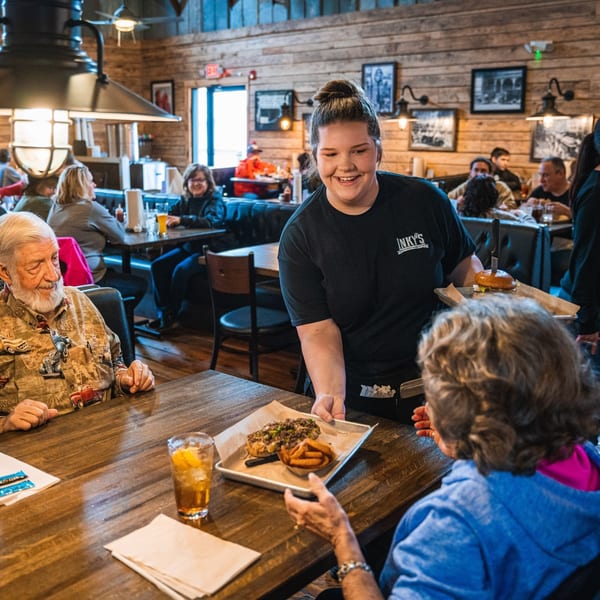 A smiling Inky's server delivers a cheesesteak and fries to a customer at a wooden table.